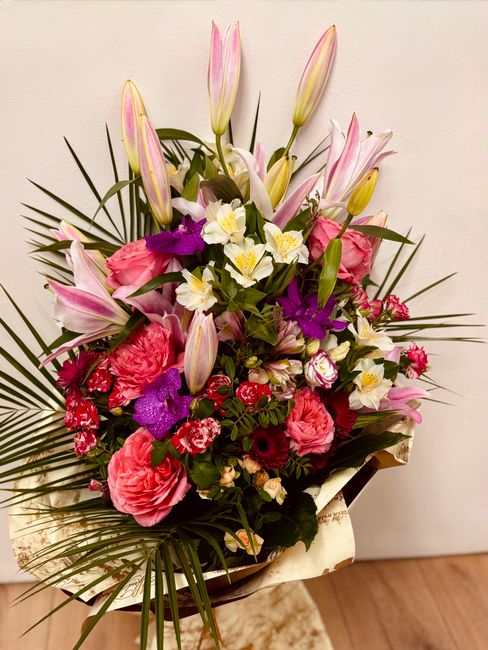A vibrant floral arrangement featuring a mix of pink lilies with closed buds and open petals, large pink roses, and purple orchids, accented with white daffodils with yellow centers. The bouquet is wr