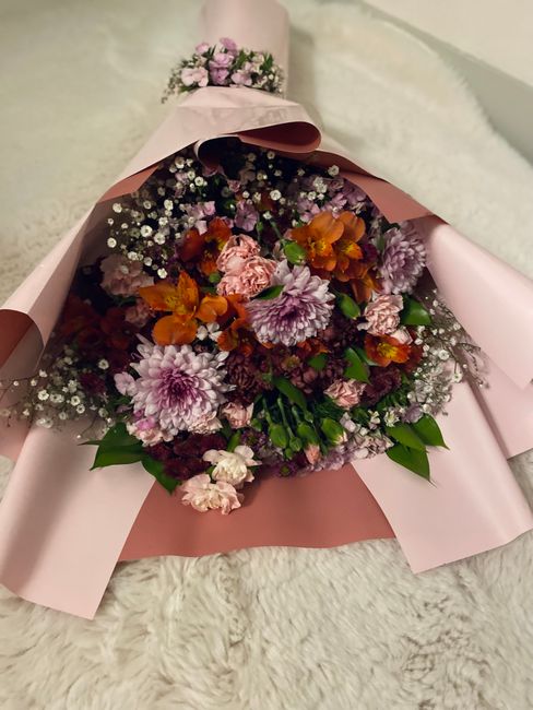 A vibrant floral bouquet featuring large, blooming pink and purple chrysanthemums, orange and pink mini carnations, and delicate white baby's breath, arranged with lush green foliage. The bouquet is w