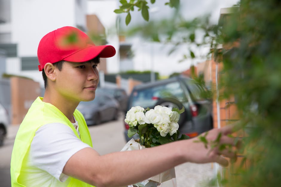 A young male florist wearing a red cap and high-visibility yellow vest is delivering a floral gift outside a residential building. He is holding a bouquet of fresh white flowers, possibly hydrangeas, 