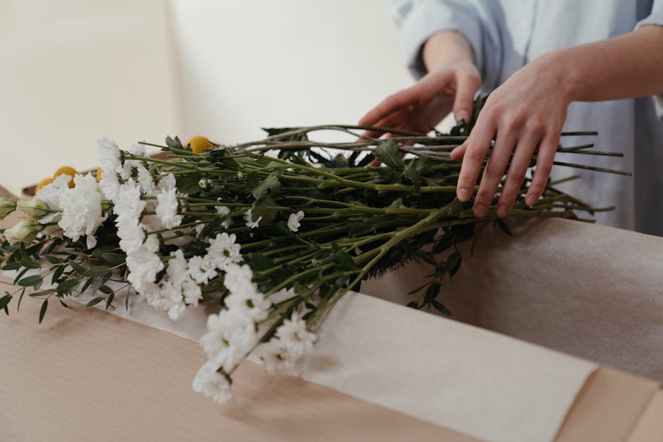 Close-up of a florist's hands carefully arranging a bouquet of fresh white carnations, yellow billy balls, and green foliage on a light wooden table. The bouquet is partially wrapped in beige craft pa