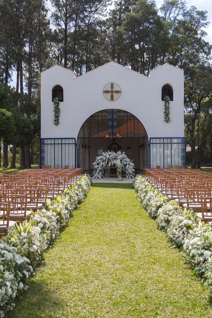 A floral arrangement featuring a large, elegant bouquet of predominantly white flowers, including roses and hydrangeas, is placed at the entrance of a white outdoor wedding chapel. The bouquet is acce