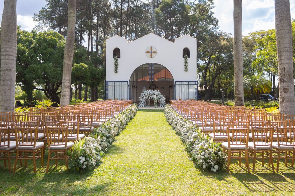 A picturesque outdoor wedding ceremony setup featuring a grassy aisle flanked by lush white floral arrangements on each side, leading to a white chapel with arched doorway and decorative cross emblem.